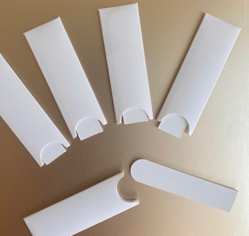 Martinenq 和 Fedrigoni 携手发现香水和化妆品 2 site-industries-cosmetiques 在浅色平面上排列着六个白色长方形纸袋。其中五个纸袋是垂直的,带有圆形开口,而标有 "Brouillon auto "字样的纸袋则是水平的,其翻盖部分从纸袋中伸出。